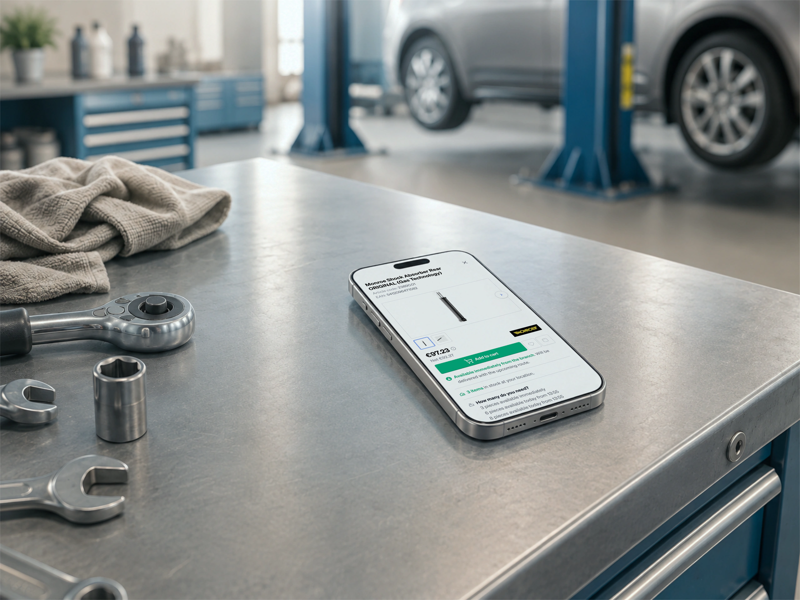 A phone on a workshop bench beside a ratchet and spanners, a shock absorber priced and ready to add to the cart on screen, a car on the lift behind