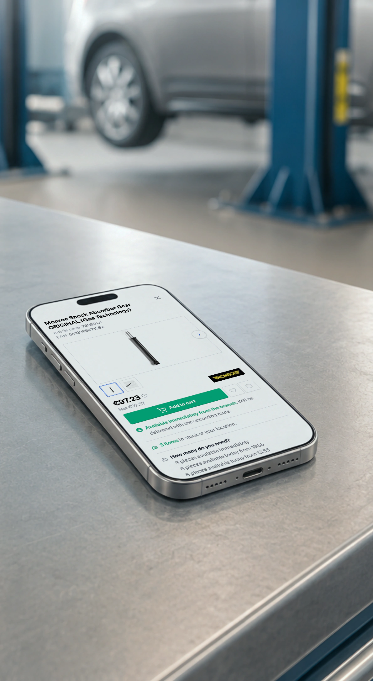 A phone on a workshop bench beside a ratchet and spanners, a shock absorber priced and ready to add to the cart on screen, a car on the lift behind